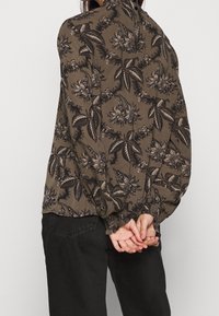 Person, die eine braune Blumenshirt mit langen Ärmeln und schwarze Hosen trägt, steht mit hinter dem Rücken gefalteten Händen vor einem hellen Hintergrund.