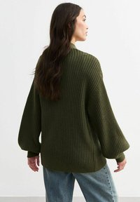Olive green oversized knitted sweater with a textured pattern, wide sleeves, and ribbed hem, paired with light blue jeans.