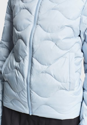 Lyseblå quiltet jakke med bølget syning, lynlås foran og sidelommer, båret over sorte bukser.