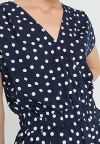 Navyblaues Kleid mit weißen Polka-Dots, ausgestattet mit einem V-Ausschnitt und einem elastischen Bund. Kurzärmel mit gerafftem Design um die Taille.