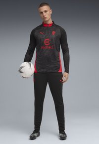 Ensemble de survêtement noir et rouge avec un haut à motifs, col zippé et logo. Pantalons noirs avec logo sur le côté, associés à des crampons noirs et verts et un ballon.