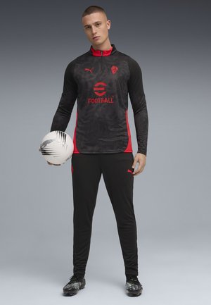 Zwarte en rode trainingsbroek met een patroon top, ritskraag en logo. Zwarte broek met zijlogo, gecombineerd met zwart en groene voetbalschoenen en een bal.