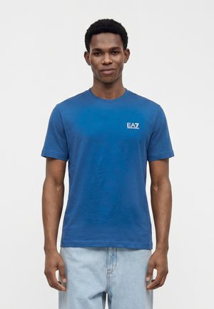 Jeune homme portant un t-shirt bleu EA7 Emporio Armani et un jean bleu clair, debout devant un fond blanc uni.