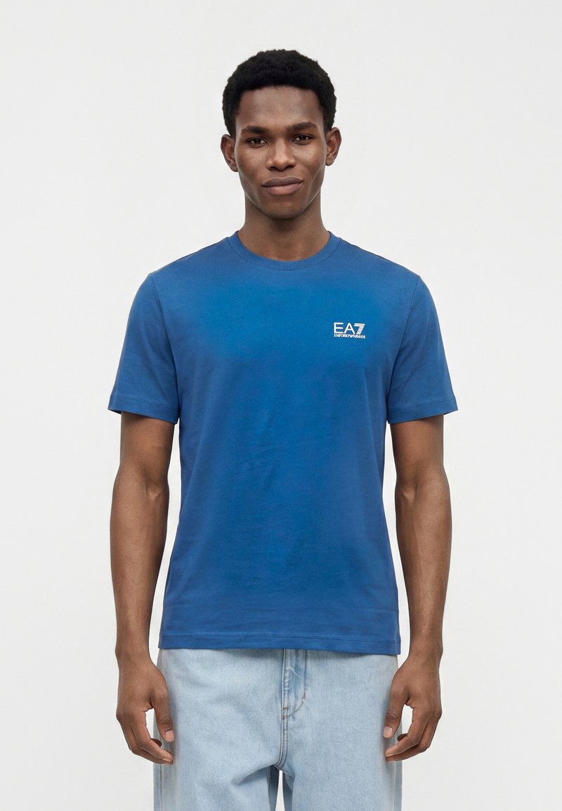 Νέος άνδρας που φοράει μπλε μπλουζάκι EA7 Emporio Armani και ανοιχτό μπλε τζιν, στέκεται μπροστά σε απλό λευκό φόντο.