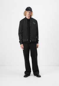 Black bomber jacket with a zip front, ribbed cuffs, and patch detail. Paired with black shirt and wide-leg trousers, finished with black shoes.