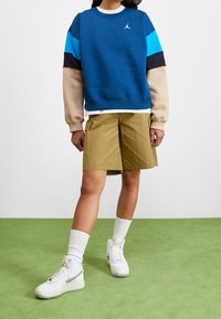 Felpa girocollo blu e beige con logo, abbinata a pantaloncini kaki. Sneakers alte bianche con un accento verde. Calzini bianchi a coste.