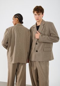 Costum două piese cu model maro și design în carouri. Include un blazer cu nasturi și buzunare, asortat cu pantaloni care se potrivesc.