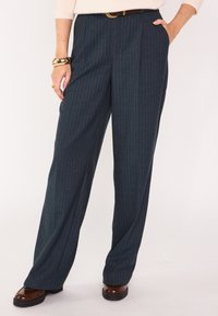 Pantalon à jambes larges en tissu bleu sarcelle foncé à fines rayures. Il présente une taille haute, des poches latérales et est associé à des chaussures marron.