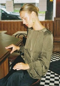 Olivgrön satinblus med puffärmar och rynkade manschetter. Modellen sitter vid ett bord, iförd stora guldörhängen, med mörka byxor.