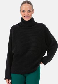 Pull à col roulé noir avec un motif côtelé texturé, coupe ample et épaules tombantes, associé à un pantalon vert.