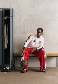 Hvit langermet skjorte med svarte og røde striper, røde bukser med logo på siden, svarte sko. Holder en brun basketball i en nettpose.