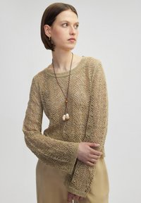 Gestrickter goldener Pullover mit einem detaillierten Spitzenmuster, weiten Ärmeln und einem Schiffsausschnitt, kombiniert mit einer langen Halskette mit dekorativen Perlen.