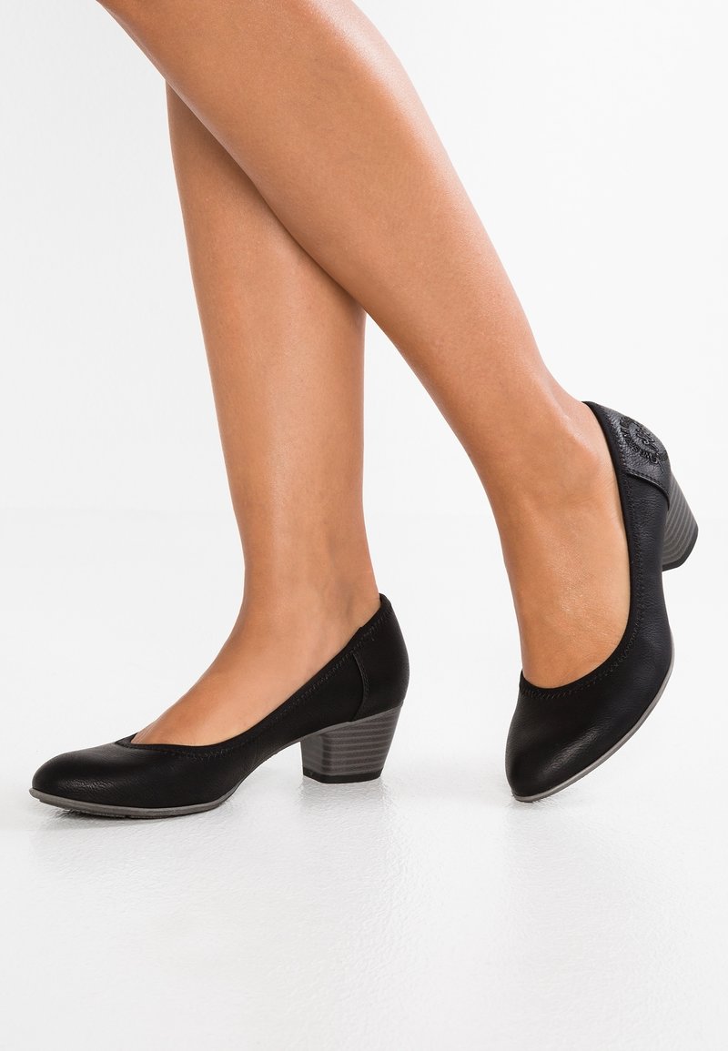 Jambes de femme portant des chaussures en cuir noir à talons bas, bouts arrondis et talons empilés, debout sur une surface blanche lisse.