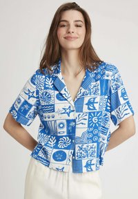 Chemise à manches courtes à boutons avec un imprimé bleu et blanc sur le thème de l'océan, intégrant divers motifs tels que des vagues, des étoiles et des dessins de coraux.