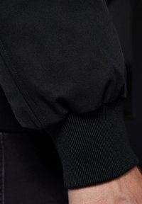 Czarny rękaw od bluzy z elastycznym mankietem, wyróżniająca się teksturowana tkanina oraz gładkie detale szwów wzdłuż szwu.