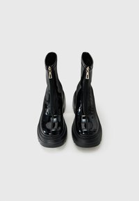 Pair of black patent leather ankle boots with front zippers and thick rubber soles, viewed from the front on a white background.