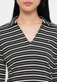 Pasiasta czarno-biała koszulka polo z dekoltem w kształcie litery V, z kołnierzykiem i małym logo po lewej stronie. Gładka, miękka faktura.