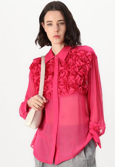 Blouse rose avec un devant texturé au motif de roses, en tissu transparent, manches longues avec poignets boutonnés, et col. Accompagnée d'un sac à main de couleur claire.