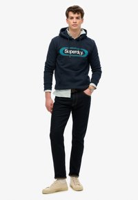 Navy bluza z kapturem z logo "Superdry", nałożona na jasną, w paski koszulę, zestawiona z ciemnymi dżinsami i beżowymi sneakersami. Luźny krój i design.
