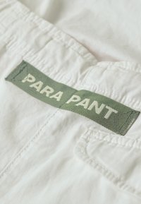 Białe spodnie z tkaniny z zieloną, tkanym metką z napisem "PARA PANT" w białym kolorze. Zawierają detale szwów i tylnią kieszeń.