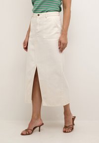 Cremfarbener Denim-Midi-Rock mit einem Schlitz vorne, zwei seitlichen Taschen und einem Knopfverschluss an der Taille. Kombiniert mit braunen Riemchenheels.