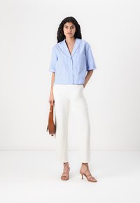 Blau-weiß gestreifte Kurzarmbluse mit Knopfleiste, kombiniert mit weißen Hosen und einer braunen Handtasche. Hellbraune Riemchenschuhe.