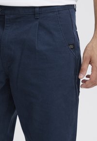 Sötétkék pamut nadrág sima textúrával, elülső zsebbel és egy kis márkajelzéssel az oldalsó varrásnál.