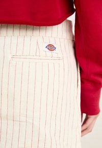Gonna crema con strisce verticali rosse, dotata di una tasca posteriore e dell'etichetta "Dickies". Il materiale sembra robusto e strutturato.