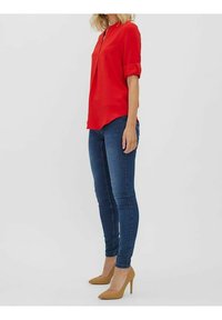 Rød, struktureret bluse med mandarinkrave; parret med tætsiddende blå jeans og beige højhælede sko. Sidevisning viser beklædningens længde og pasform.
