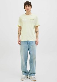 Lichtgele katoenen T-shirt met een relaxte pasvorm en geprinte tekst "ORIGINALS". Gekocht met vervaagde blauwe, losse jeans en grijze sneakers.