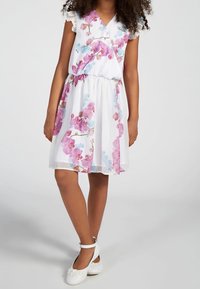 Pige iført en hvid knælang kjole med lyserøde og blå blomsterprints samt hvide ankelsandaler med blomsterudsmykninger.