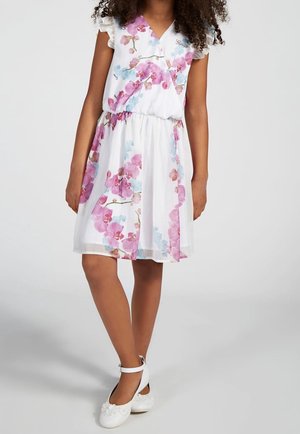 Ragazza in un vestito bianco fino al ginocchio con stampe floreali rosa e blu e scarpe bianche con cinturino alla caviglia decorate con fiori.