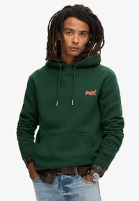 Felpa verde scuro con tasca frontale, cordini regolabili e piccolo logo ricamato. Camicia a quadri con colletto visibile sotto.