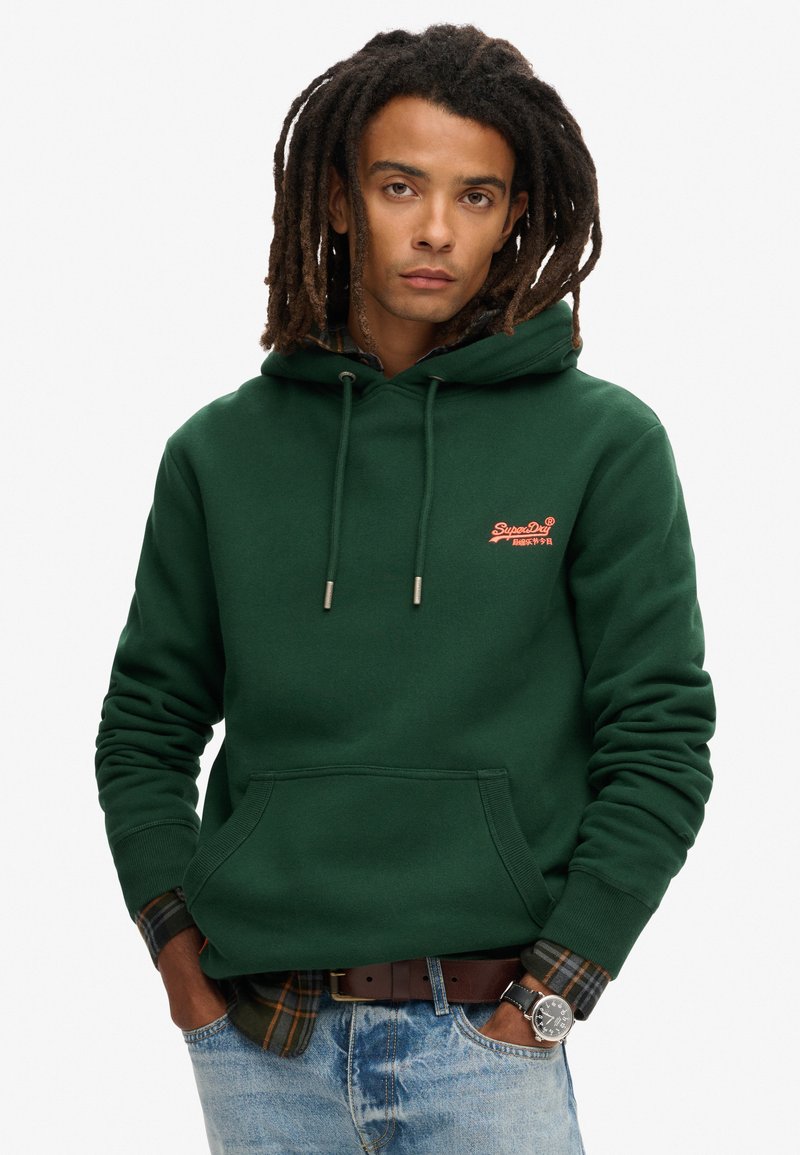 Felpa verde scuro con tasca frontale, cordini regolabili e piccolo logo ricamato. Camicia a quadri con colletto visibile sotto.