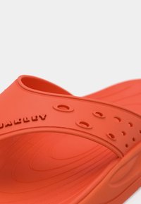 Πορτοκαλί πλαστική σαγιονάρα με φαρδύ λουρί με κοψίματα, υγρό πέλμα και ανάγλυφο όνομα μάρκας στο λουρί.