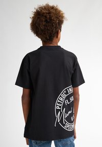Sort bomulds T-shirt med korte ærmer, med hvidt cirkulært logo trykt på ryggen. Enkel design med en klassisk pasform.