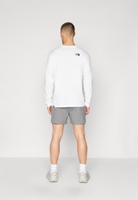 Balta ilgomis rankovėmis marškiniai su juodu logotipu ant nugaros, derinama su pilkomis šortais ir pilkomis sportinėmis avalynėmis. Modelis atsisukęs atgal, pristato aprangą.