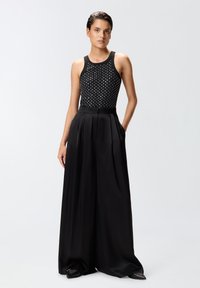 Body nero senza maniche con applicazioni di borchie argentate, abbinato a pantaloni neri in raso plissettati a vita alta. Texture liscia, silhouette elegante.