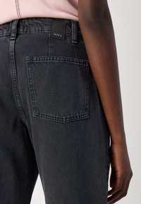 Svarte denimbukser med en rektangulær baklomme og en liten svart merkeetikett i livet. Stoffet har en teksturert overflate.