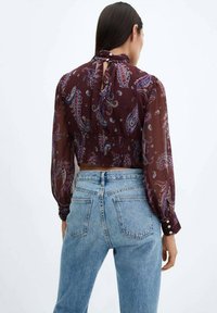 Blouse en soie à motif paisley bordeaux avec de longues manches bouffantes, détail en goutte d'eau dans le dos et taille cintrée, associée à un jean en denim bleu taille haute.