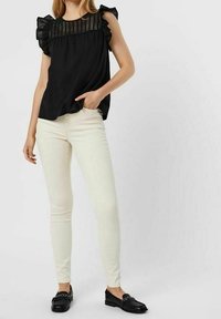 Donna vestita con un top senza maniche nero a balze, jeans skinny bianchi e mocassini neri con dettaglio a catena, in piedi con una mano in tasca.