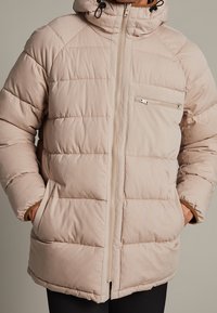 Ljus beige puffjacka med dragkedja fram, huva och snedstickning; har en dragkedjeförsedd bröstficka och elastiska ärmslut.