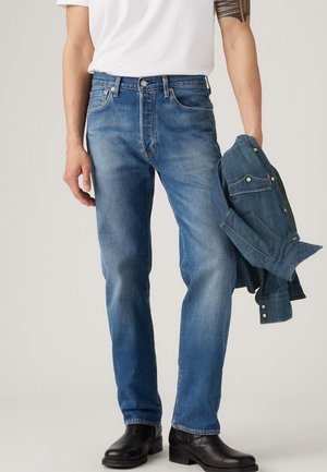 Homme portant un jean bleu, des bottes noires et un t-shirt blanc tenant une veste en denim bleu pliée dans sa main gauche.