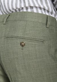 Primer plano de bolsillo de pantalones ajustados verde oliva con detalle de botón, usados con una camisa a rayas metida dentro.