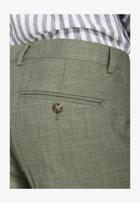 Primer plano de bolsillo de pantalones ajustados verde oliva con detalle de botón, usados con una camisa a rayas metida dentro.