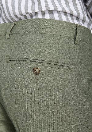 Nahaufnahme einer olivgrünen, maßgeschneiderten Hosentasche mit Knopfdetail, getragen zu einem gestreiften, hineingesteckten Hemd.