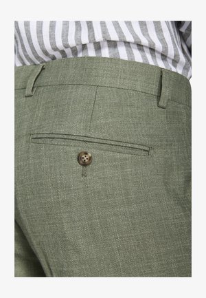 Nahaufnahme einer olivgrünen, maßgeschneiderten Hosentasche mit Knopfdetail, getragen zu einem gestreiften, hineingesteckten Hemd.