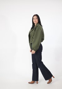 Oliwkowa krótka kurtka z dużymi rękawami i złotymi guzikami, zestawiona z ciemnymi rozszerzanymi dżinsami i brązowymi butami na obcasie.