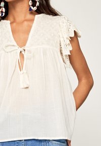 Witte, lichte blouse met geborduurde schouderpartij, striksluiting, kwastaccent en kant met ruffles aan de mouwen. Heeft een relaxte pasvorm en een brede halslijn.