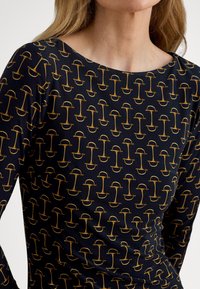 Svart langermet skjorte med et gyllent geometrisk mønster. Har en scoop-utrigning og en tettsittende silhuett. Mykt, elastisk stoff.